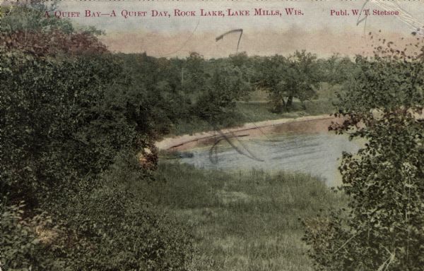 View down hill towards the curving shoreline of Rock Lake, surrounded by trees and long grass. Caption reads: "A Quiet Bay — A Quiet Day, Rock Lake, Lake Mills, Wis."