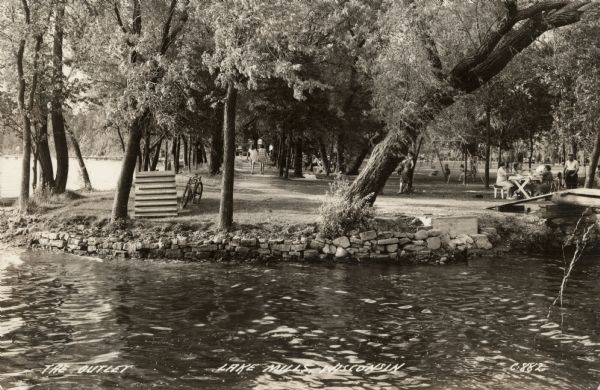 View across outlet towards the park by Rock Lake. People are picnicking, children are on a swing set, and women are walking along a path. There is a rustic wood bridge on the far right over the outlet. Caption reads: "The Outlet, Lake Mills, Wisconsin."