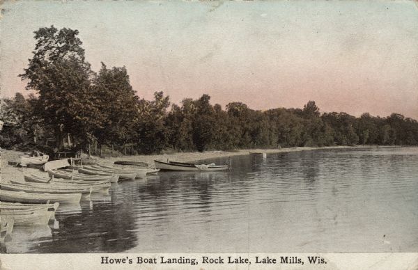 View across water towards a row of rowboats lined up on the shoreline of Rock Lake. Caption reads: "Howe's Boat Landing, Rock Lake, Lake Mills, Wis."
