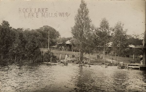 View across water towards the shoreline of Rock Lake. People are in rowboats at the shoreline. There are cabins behind the trees, and on the right is a pier and boathouse over the water. Caption reads: "Rock Lake, Lake Mills, Wis."