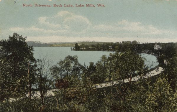 Elevated view through trees of a road along the shore of Rock Lake. A large dwelling is further down the road on the right. Caption reads: 
"North Driveway, Rock Lake, Lake Mills, Wis."