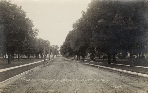 View down unpaved tree-lined street in a residential neighborhood. A park is on the left, and houses are on the right. Caption reads: "Madison St. looking West, Lake Mills, Wis."