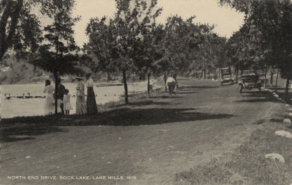 View across dirt road along a lake towards a group of people on the edge of the road looking at the lake. Automobiles are parked on the right along the curve in the road. A man is walking down the road with a bicycle near where boats are pulled up along the shoreline near a pier. Caption reads: "North End Drive, Rock Lake, Lake Mills, Wis."