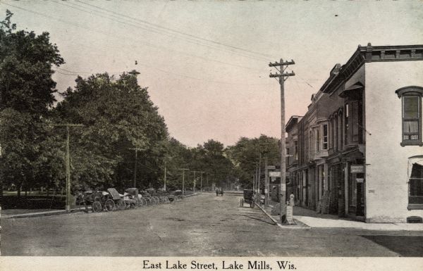 View down street towards a row of buggies parked on the left along a park, across from commercial buildings on the right. Caption reads: "East Lake Street, Lake Mills, Wis."