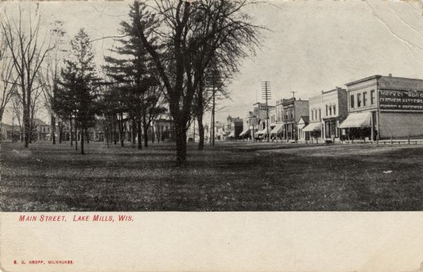 View from park towards the central business district. Businesses include a tailor's shop and hardware store. Caption reads: "Main Street, Lake Mills, Wis."