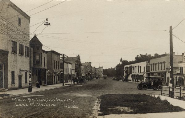 View down street towards the central business district. Horses, buggies and automobiles are in the street. Caption reads: "Main St. Looking North, Lake Mills, Wis."