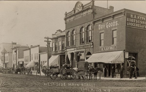 View across unpaved street towards a city block, (Williams Block) lined with businesses. They include: H.A. Kypke Dry Goods, Heimstreets Drug Store, and a saloon. Horses and wagons are parked at the curb. A group of men are standing at the street corner in front of the H.A. Kypke storefront. Caption reads: "West Side Main Street, Lake Mills, Wis."