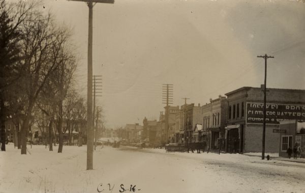 Downtown Lake Mills after a snowfall. A park and residential block are on the left. Commercial businesses are on the right. Horses and wagons are parked in the street.