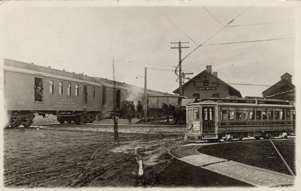 View towards the Lake Mills Railroad Depot. A passenger train is on the tracks on the left. A light rail trolley is on the sharply curving tracks on the right.