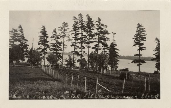 View of Lake Nebagamon from across a fenced-in garden. Automobiles are parked along the lakeside road.