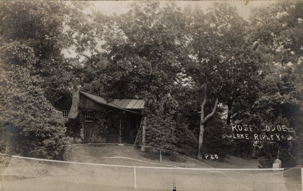 View towards a cottage nestled among the trees. In the foreground a tennis net is stretched across the sloped lawn. Caption reads: "Rose Lodge — Lake Ripley."