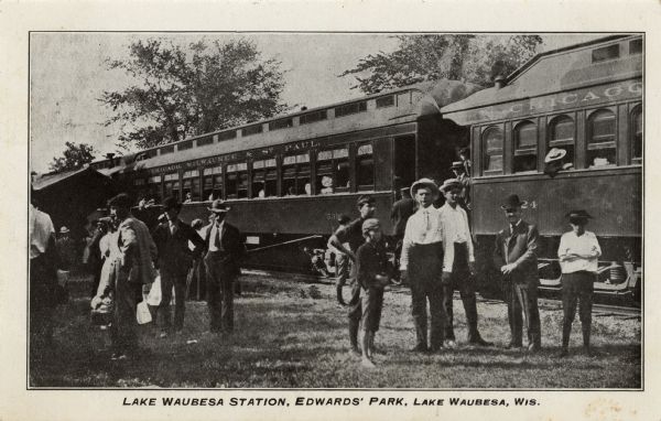 View of Chicago, Milwaukee & St. Paul passenger cars stopped behind a group of men and boys standing in the foreground. Caption reads: "Lake Waubesa Station, Edwards' Park, Lake Waubesa, Wis."