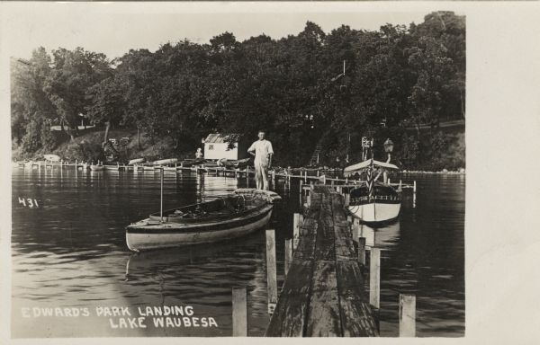 View down a dock with two boats on either side. A man is standing on the one on the left. There is a boathouse on the lakeshore, with several canoes and rowboats tied up along a long pier. Caption reads: "Edwards' Park Landing, Lake Waubesa, Wis."