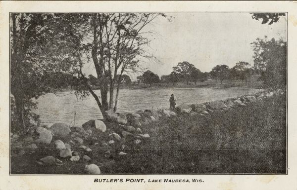 View of part of a lake with boulders along the shore. A man is is standing among the rocks at the shoreline. Caption reads: "Butler's Point, Lake Waubesa, Wis."