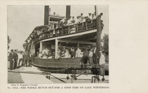 View of a double-deck excursion boat filled with a crowd of tourists. A few men are standing on the sidewalk to the side.