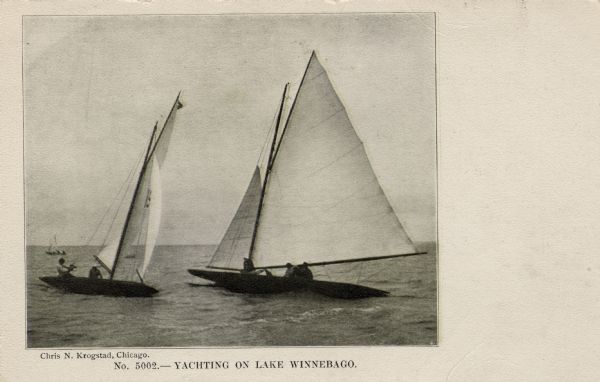 View across water towards a crew of men racing two sailboats. Another boat is in the background on the left.