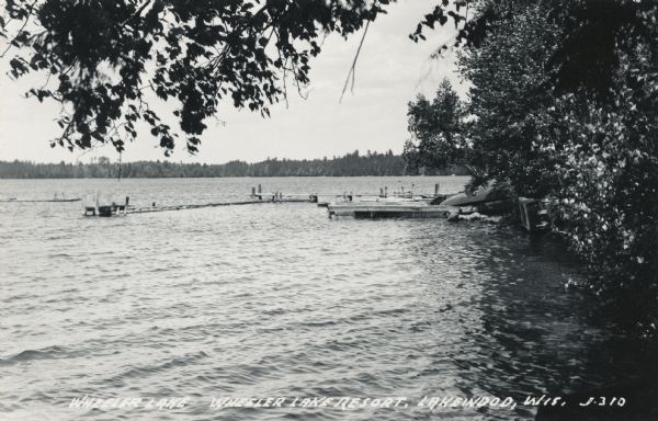 View across water along shoreline towards a long dock and a raft. A rowboat is turned over on the dock near the tree-lined shoreline. Caption reads: "Wheeler Lake, Wheeler Lake Resort, Lakewood, Wis."