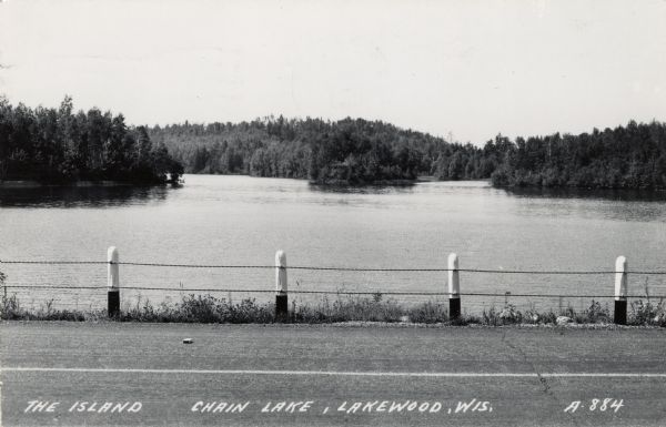 View across paved road with a guard fence towards an island in a lake.