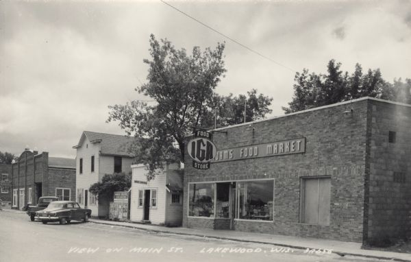 Photographic postcard view of a central business block, featuring Adams Food Market IGA and a Post Office. To the left of the Post Office is a signboard for Wabeno Theatre that includes show posters. Gilbert's Bar is further down on the left. A car and a flatbed truck are parked at an angle at the curb. Caption reads: "View on Main St., Lakewood, Wis."