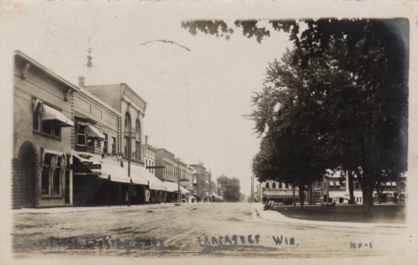 View down unpaved town street. Businesses include a dentists' office, Lathrop's, Joeckel Bros., and Hatch Drugs on the left. There is a park on the right, with people sitting on benches on the sidewalk. Caption reads: "Maple St., Looking East, Lancaster, Wis."