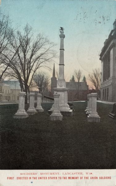 The Soldiers' Monument, first erected in the United States to the memory of the Union Soldiers. In the center on a lawn is a tall marble column with Old Abe (eagle symbol) at the top holding a wreath, perched on an orb. The monument is surrounded by smaller marble columns and a cannon. The courthouse is on the right, and a church building is in the far background. Caption reads: "Soldiers' Monument, Lancaster, Wis. First erected in the United States to the memory of the Union Soldiers."
