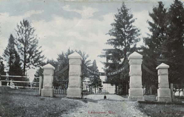 View of the entrance gate and stone columns for a road to a cemetery. Caption reads: "Hillside Cemetery, Lancaster, Wis."