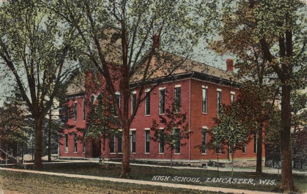 View across unpaved road towards a two-story red brick school building. Caption reads: "High School, Lancaster, Wis."