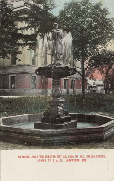 Hand-colored postcard of the Memorial Fountain, with the Grant County Courthouse in the background. Caption reads: "Memorial Fountain erected May 30, 1906, by McKinley Circle, Ladies of G. A. R., Lancaster, Wis."