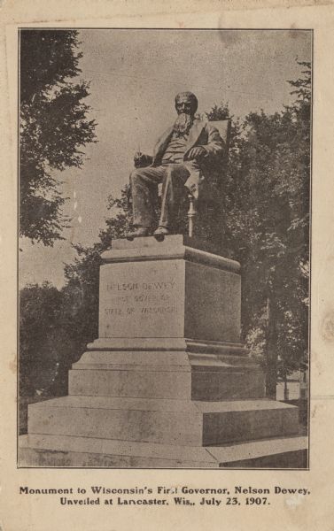 A monument to Nelson Dewey featuring a sculpture of him seated on a chair on top of a pedestal. He is holding a pen in his right hand. Caption reads: "Monument to Wisconsin's First Governor, Nelson Dewey. Unveiled at Lancaster, Wis., July 23, 1907."