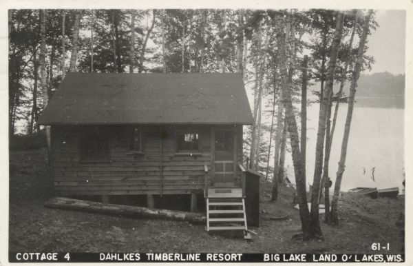 View down hill towards a small guest cottage surrounded by birch and other types of trees next to Big Lake at Dahlkes Timberline Resort. Rowboats are on the shoreline near the dock further down the hill on the right. Caption reads: "Cottage 4, Dahlkes Timberline Resort, Big Lake, Land O' Lakes, Wis."