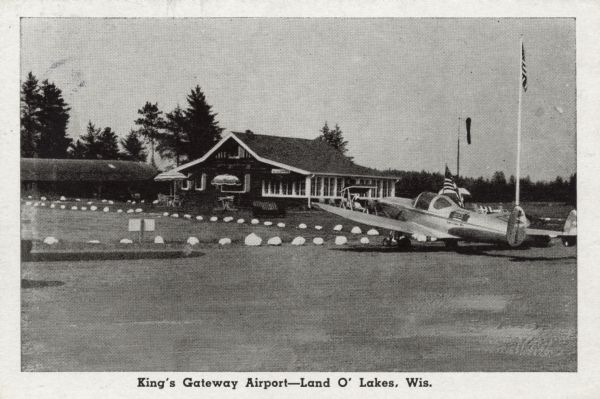 View towards a small airport with seating under umbrella's on a patio. A small plane is parked on the tarmac in the right foreground. Caption reads: "King's Gateway Airport — Land O' Lakes, Wis."