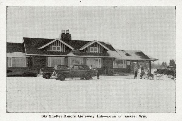 View towards a ski lodge. People with dogs are gathered near automobiles parked in front. Caption reads: "Ski Shelter King's Gateway Hill — Land O' Lakes, Wis."