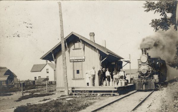 View across tracks towards a small railroad depot, with people gathered on the platform. A locomotive belching smoke is approaching the depot.
