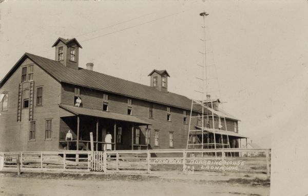 View of a large boarding house. Two people are standing on the porch, and a woman is sitting and looking out of the sill of a second-story window. Caption reads: "Company Boarding House, Laona, Wis."