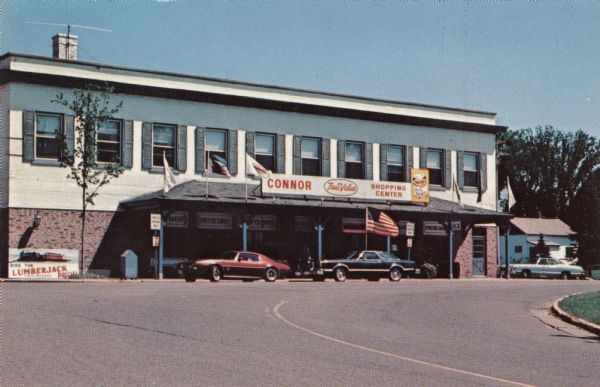 Color postcard of a series of shops in a single building, including: a bakery, a coffee shop, a True Value Hardware Store, a gift shop and a sporting goods store. Automobiles are parked in front.