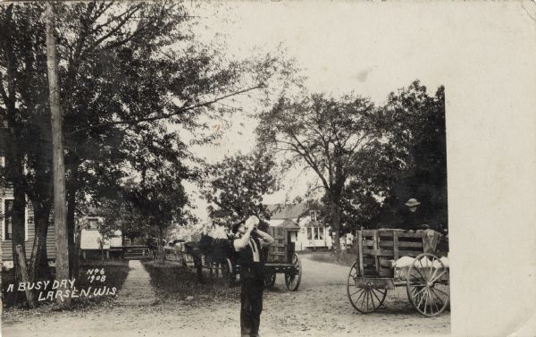 A man is standing in the street drinking from a jug, and a woman sitting in a wagon is watching him. Horses and wagons are behind him. Caption reads: "A Busy Day, Larsen, Wis."