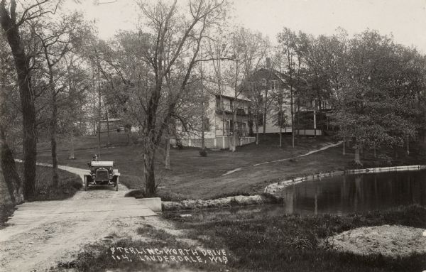 View of a man driving an automobile down a road, approaching a wooden bridge over a stream next to a pond. Dwellings are on the slope of a hill in the background. Caption reads: "Sterlingworth Drive, 104, Lauderdale, Wis."