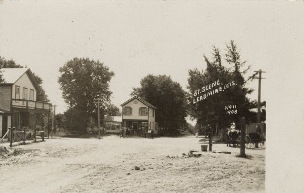 View down unpaved street with a few wooden commercial buildings. The building in the center background has a group of people gathered in front, and along the wall on the left are advertisements for a fair. There is a water pump in the foreground on the right and a horse-drawn vehicle is behind it. Caption reads: "St. Scene, Leadmine, Wis."