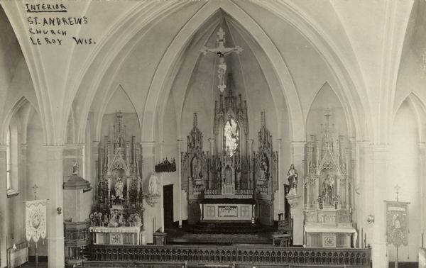Photographic postcard of an elevated view of an ornate altar with religious statuary and Gothic arches. A crucifix is hanging from the top of the arch over the altar. Caption reads: "Interior, St. Andrews Church, LeRoy, Wis."