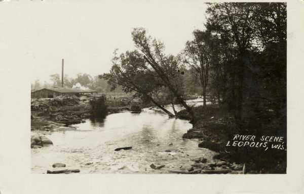 View of a small river with logs piled on the left bank near an industrial building with a smokestack. Caption reads: "River Scene, Leopolis, Wis."