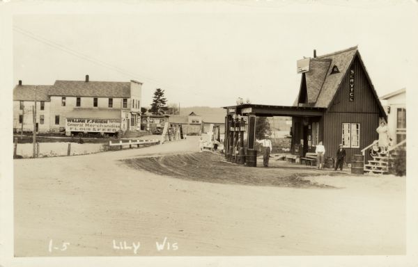 Photographic postcard view of the road through a small settlement. A dwelling and a service station are on the right. A general store on the left is on the other side of a bridge crossing a river. A man, a woman and two children are standing near the service station. Three children are standing near the bridge guardrails in the center. Caption reads: "Lily, Wis."