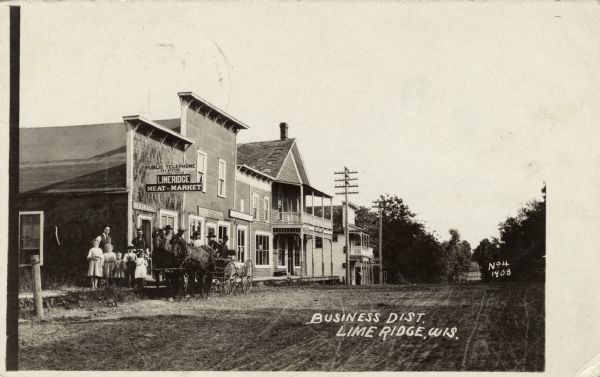 View down unpaved road going through a settlement. A meat market with a public telephone is on the left, with a group of people gathered on the sidewalk in front. A horse and buggy is along the curb. Caption reads: "Business Dist., Lime Ridge, Wis."