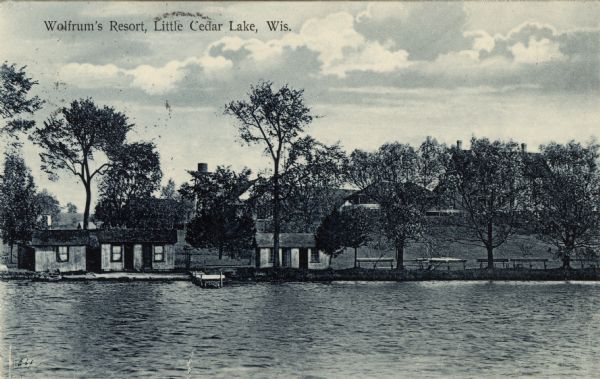 View across lake towards three guest cabins under trees on the shoreline near a pier. The main building is on the hill behind. Caption reads: "Wolfrum's Resort, Little Cedar Lake, Wis."