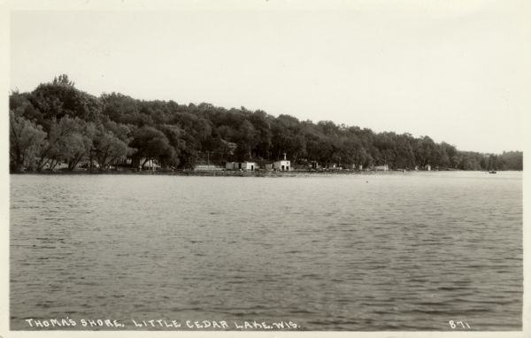 Photographic postcard view across lake towards boathouses along a lake shore.  Caption reads: "Thoma's Shore, Little Cedar Lake, Wis."