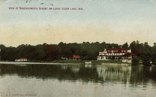 View across water towards a lakeside resort. The largest building has three stories, and below it is a small, sandy beach near a dock. Caption reads: "View of Rosenheimer's Resort on Large Cedar Lake, Wis."