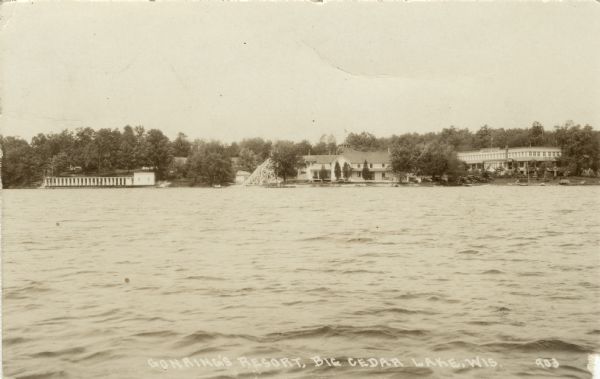 View across water towards a hotel on the lake. There is a large slide at the shoreline in the center, and near a long building on the left is what may be a diving platform. Caption reads: "Gonring's Resort, Big Cedar Lake, Wis."