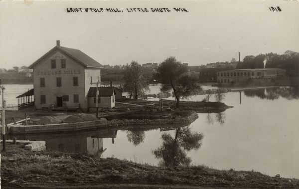 Zeeland Mills on the Fox River. Little Chute is across the river on the hill. A bridge and a dam are in the river, with industrial buildings on the far shore. Caption reads: "Grist & Pulp Mill, Little Chute, Wis."