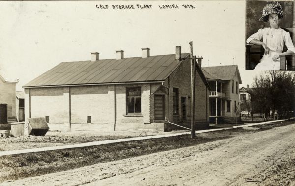 View across unpaved road towards a brick warehouse next to a dwelling. Attached to the postcard is an inset portrait of a seated women in the upper right corner. Caption reads: "Cold Storage Plant, Lomira, Wis."