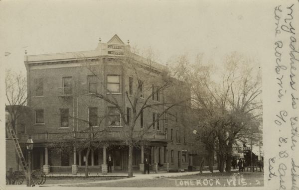 View down street towards the Dickerson House, with a sign above the entrance along the rooftop. There is a billiard hall and saloon further down the block. Caption reads: "Lone Rock, Wis."
