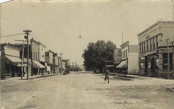 View of a main street. Meat markets and a saloon are on the left, and hardware stores and a billiard hall are on the right. A railroad crossing is in the distance. "Caption reads: "Main Street, Looking South, Lone Rock, Wis."